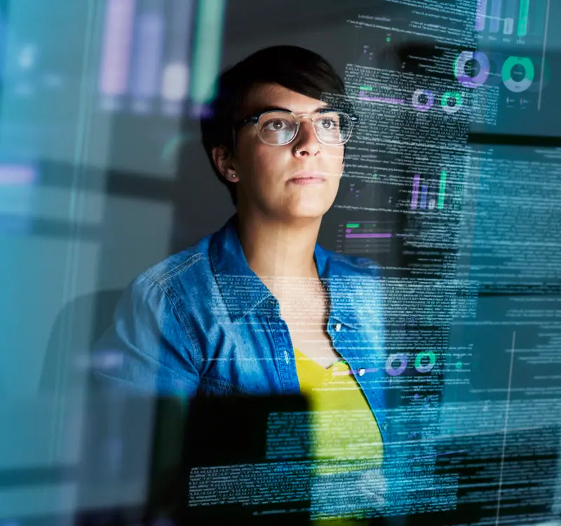 Focused woman wearing glasses working on computer with data visualizations reflected on the screen.